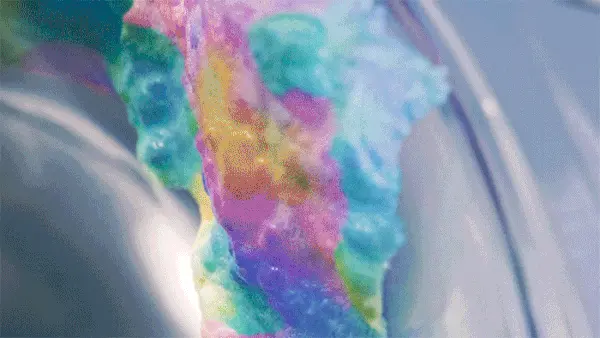 Iridescent fluid forms in blue, pink and green flowing across a pale surface.